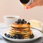 pouring maple syrup on a stack of blueberry lemon ricotta pancakes