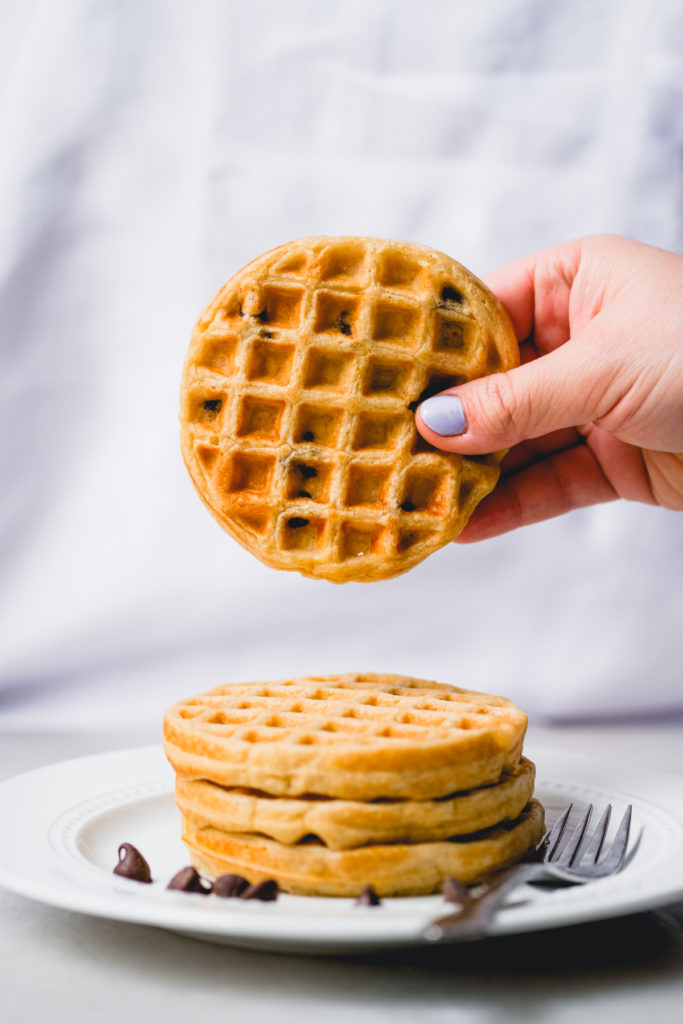 holding a mini chocolate chip oat waffle above a stack on a plate