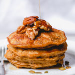 short stack of carrot cake pancakes with nuts on top with a drizzle of maple syrup